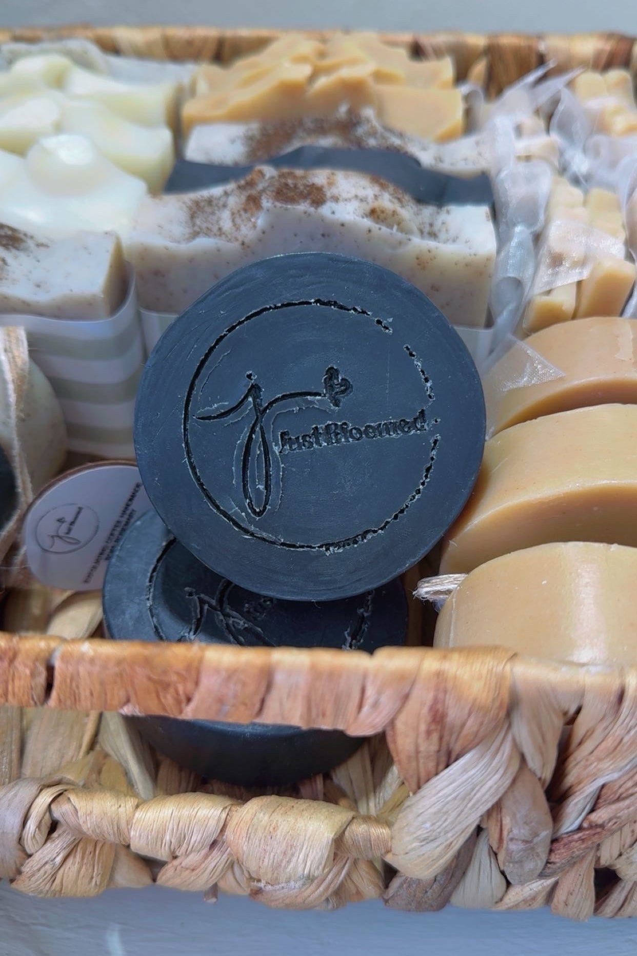 Basket of assorted soaps with a black soap bar featuring the Just Bloomed logo.